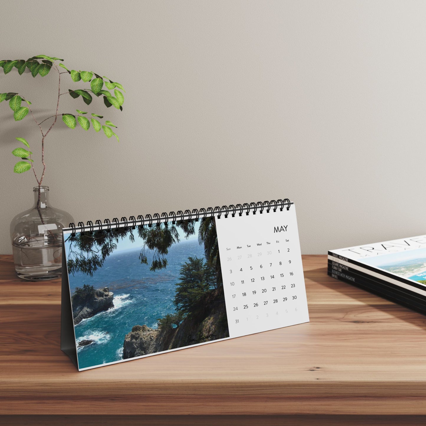 The Printify 2026 Scenic-Landscapes Desktop Calendar, open to May and showcasing a beautiful coastal photo, sits on a wooden desk next to a glass vase with a small plant and two closed magazines.