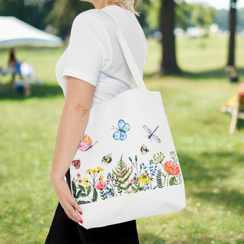 A person in a white shirt holds Printify's Spring Garden Tote Bag, showcasing colorful flowers, bees, a butterfly, a dragonfly, and a ladybug.