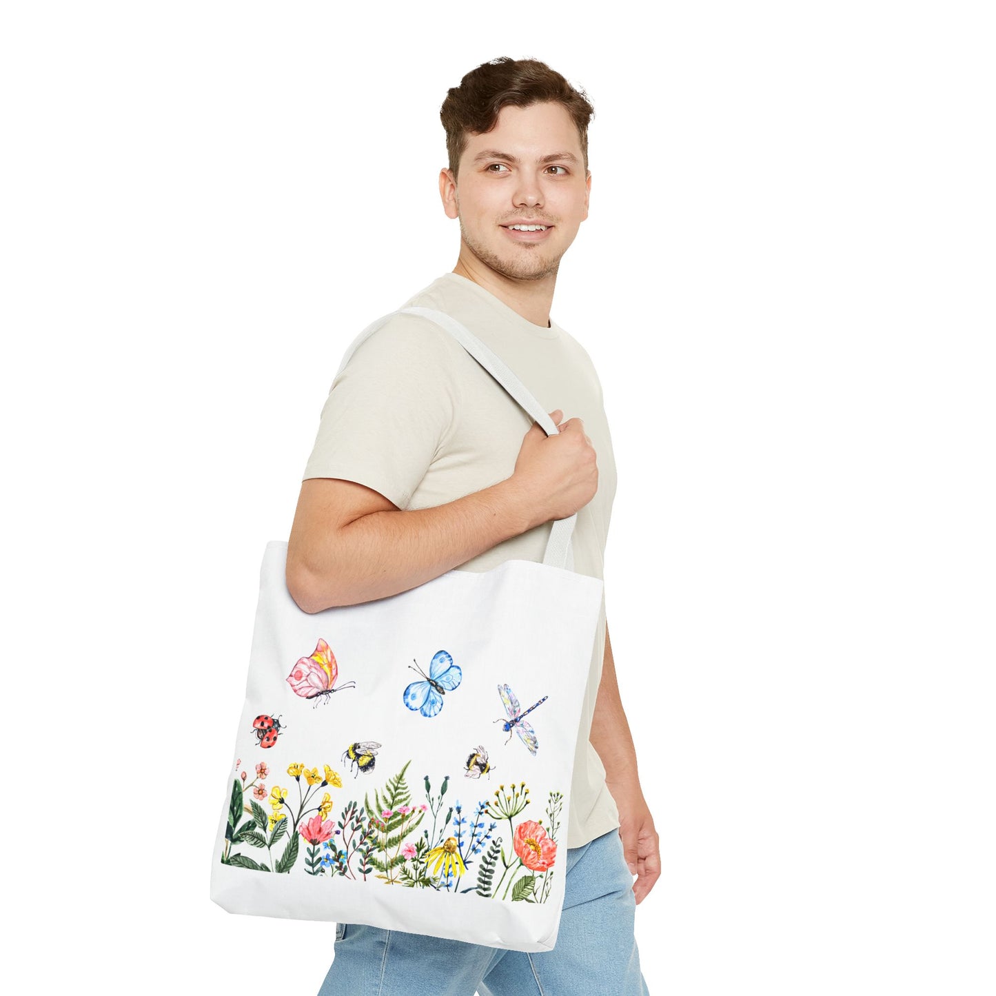 A man beams while holding his Printify Spring Garden Tote Bag, adorned with colorful flowers and insects. Clad in a neutral t-shirt and jeans, he stands against a white backdrop, prepared for outdoor adventures.