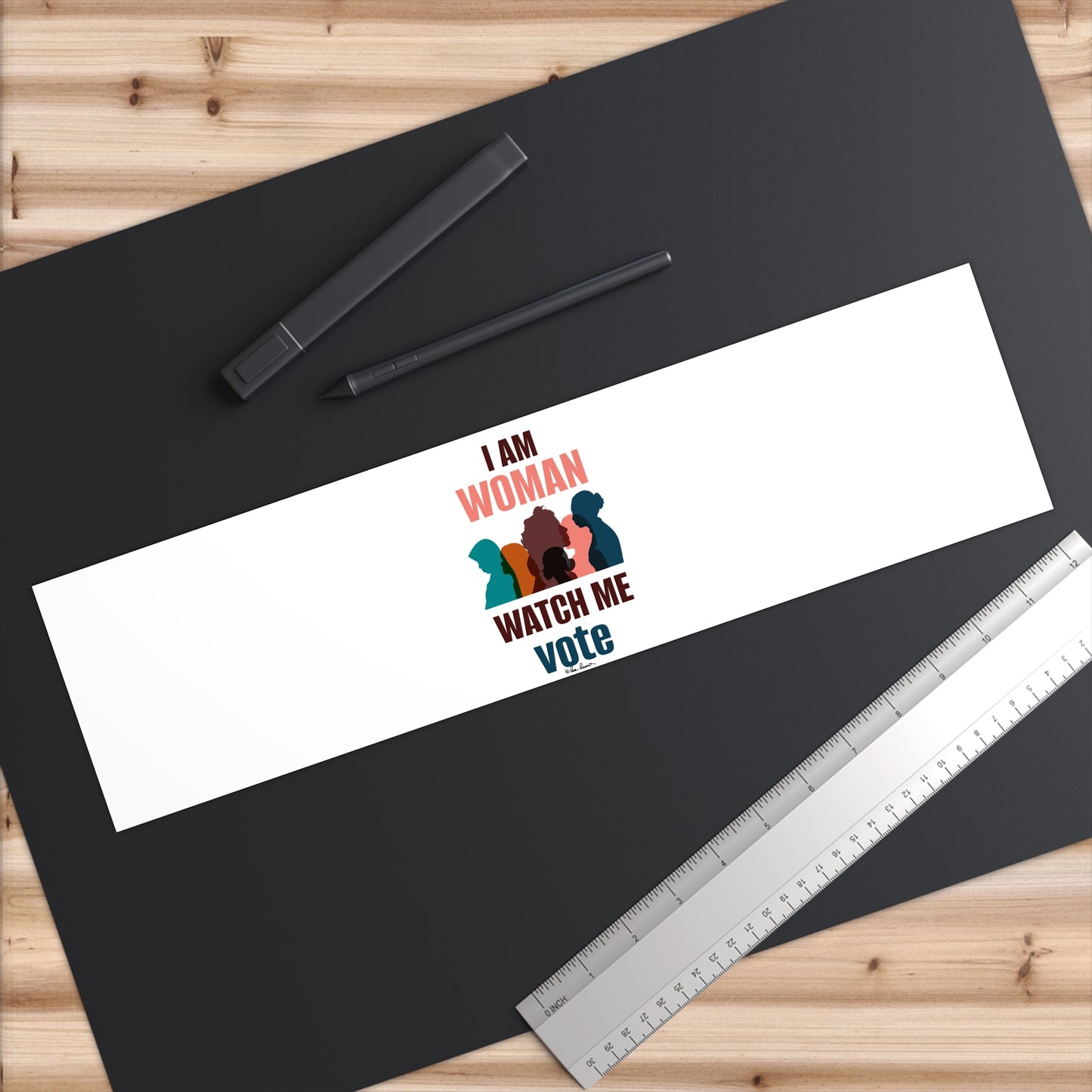 A black paper with a white Printify Voting Women's Bumper Sticker made of premium water-resistant vinyl, surrounded by two pens and a ruler on a wooden surface.