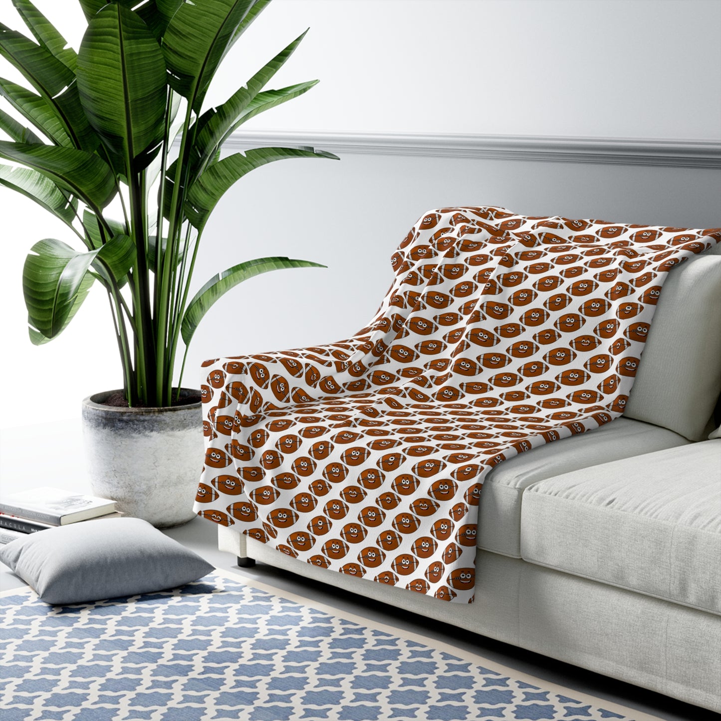 A living room with a light gray sofa adorned with Printify's Happy Football-Faces Blanket, available in 2 sizes, made from Sherpa and polyester, and featuring a cheerful brown emoji pattern. A gray pillow rests on a blue and white patterned rug, and nearby, a large potted plant adds a touch of green—perfect for sports enthusiasts to relax in style.