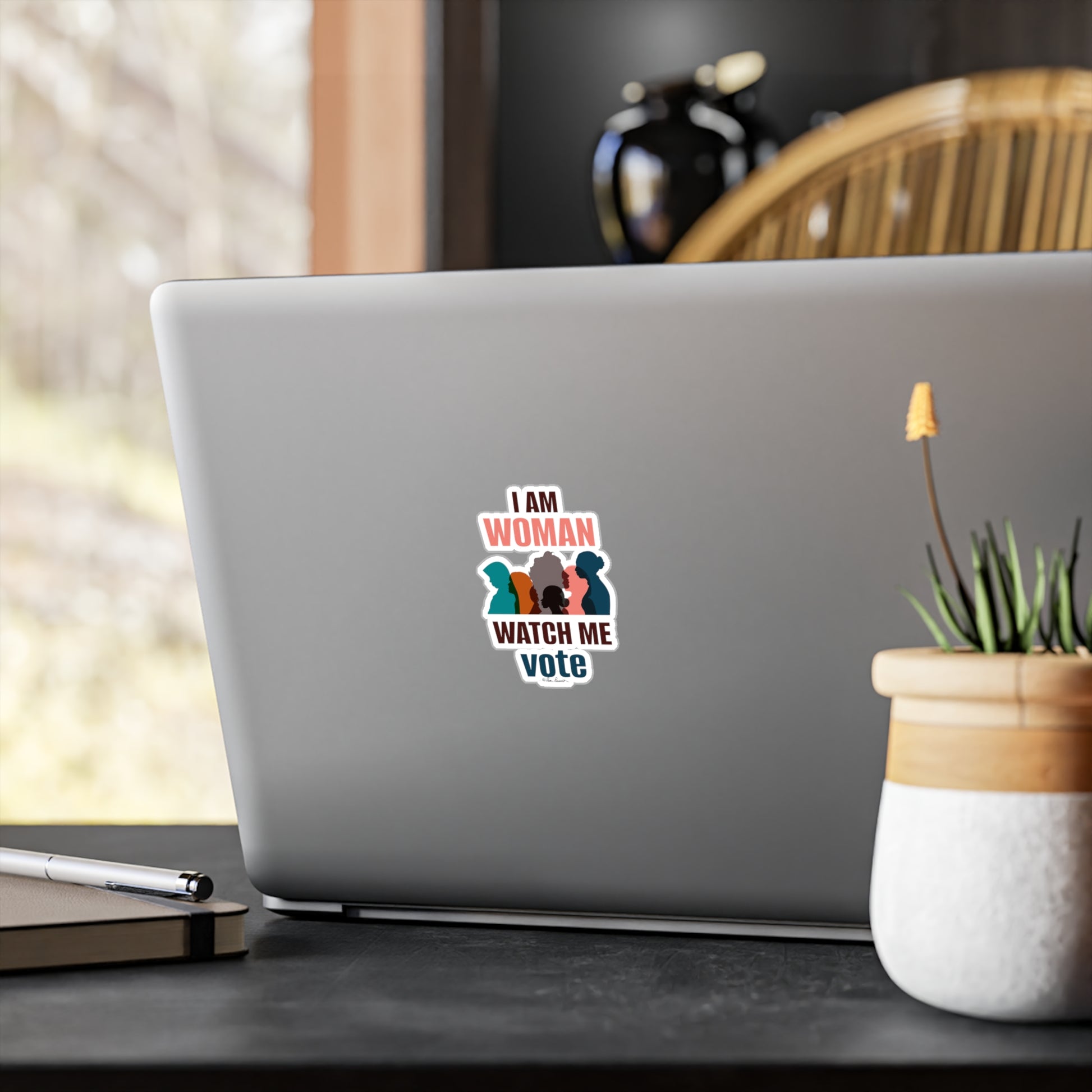 A laptop on a wooden desk with a Printify Voting Women's Decals sticker saying "i am woman watch me vote" featuring three silhouetted figures.