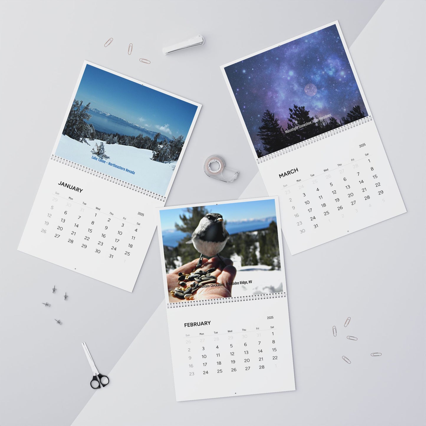 Three pages from the Minimalist 2025 Wall Calendar by Printify, showcasing January's snowy landscape, February's bird on a hand, and March's starry night sky, are scattered on a desk. Nearby are scissors, tape, and paperclips.