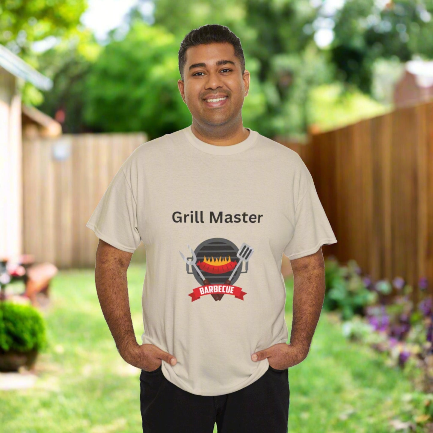 A person stands outside in a backyard, smiling and wearing a beige Printify Unisex Grill-Master T-shirt: 2 colors; Cotton; Gildan that says "Made in America" with a barbecue graphic.