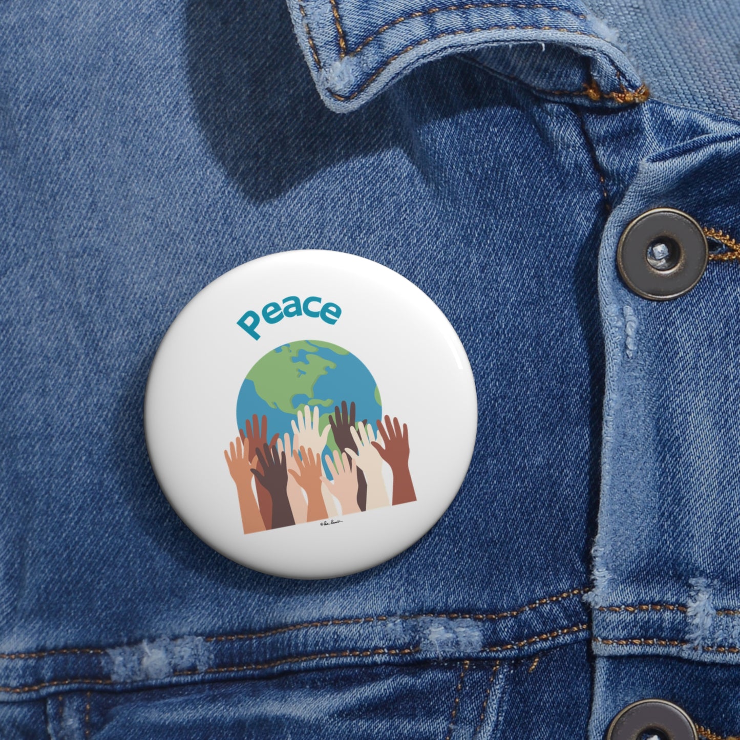 A round, American-made button from Printify featuring an image of hands of various skin tones reaching up to a globe with the word "Peace" above. This unisex World-Peace button, available in 3 sizes and made of lightweight metal, is pinned to a blue denim jacket.