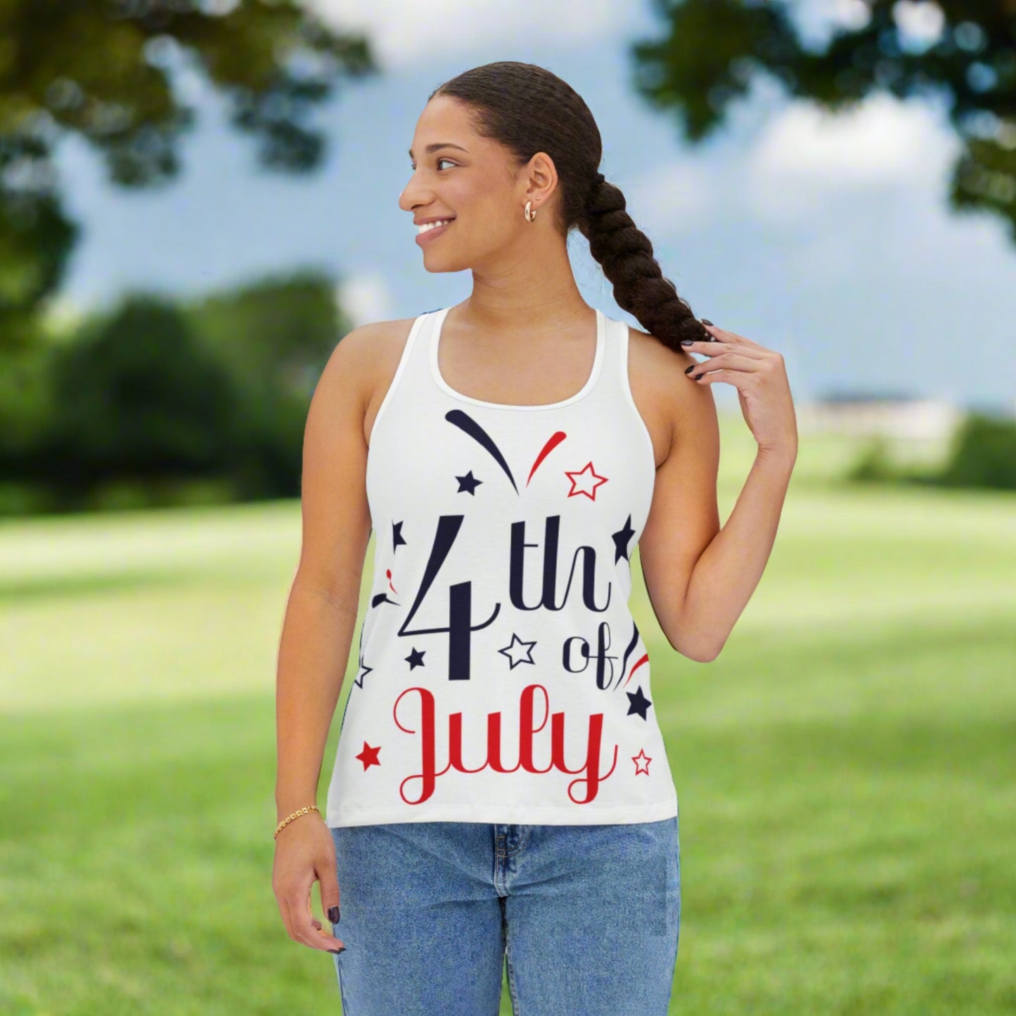 A person wearing an "Assembled in the USA" 4th of July Tank-top: Women's; Festive; Polyester blend by Printify stands outdoors, holding their braid and smiling, with green trees and a cloudy sky in the background.