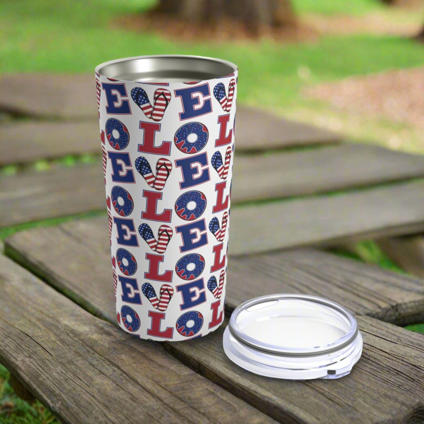 A white Printify Love USA Tumbler: 20 oz.; Stainless steel; Insulated with various 'LOVE' prints, where the 'O' is designed as a donut, stands on a wooden surface outdoors. Made of stainless steel and vacuum insulated, it keeps your drinks at the perfect temperature. The lid is placed beside it.