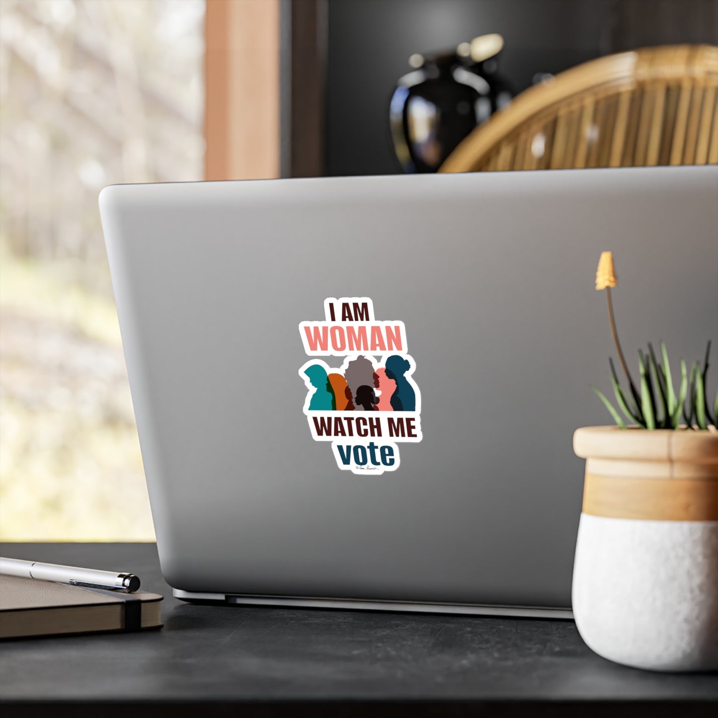 Replace with: Laptop with a Printify brand Voting Women's Decals sticker reading "i am woman watch me vote," featuring white vinyl silhouettes of three women, placed on a desk near a window.