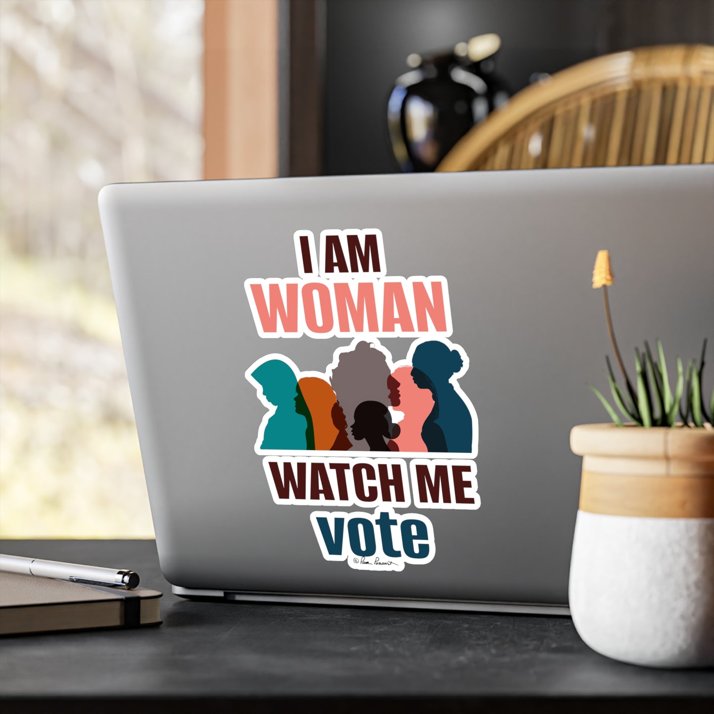 A laptop with a white Voting Women's Decals sticker by Printify featuring silhouettes of women saying "i am woman watch me vote" placed on a desk near a window.