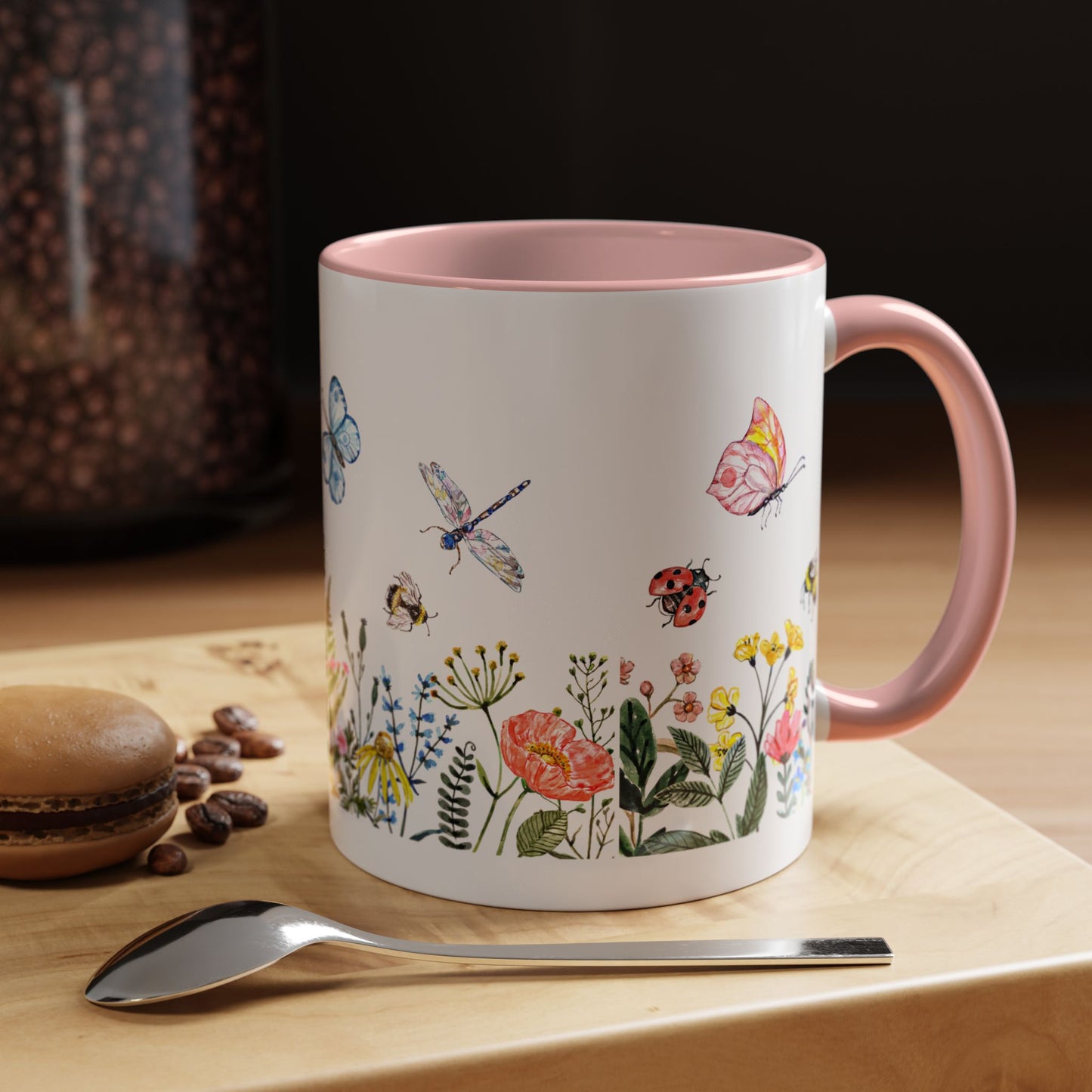 The Printify Colorful Spring Garden Mug is ideal for garden lovers, featuring a vibrant floral AOP design with a white exterior, pink interior, and handle. It complements any wooden surface beside coffee beans, a spoon, and a macaron.