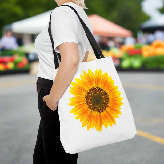 At a lively market, someone holds a white Printify Sunflower Tote Bag featuring a large, vibrant sunflower print on both sides. Blurred stalls enhance the sunny atmosphere.