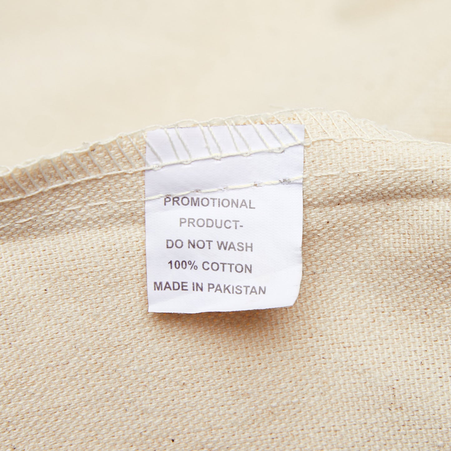 Close-up of a white clothing label on a beige fabric, with text: "PROMOTIONAL PRODUCT- DO NOT WASH 100% COTTON MADE IN PAKISTAN" displayed on a Printify Unisex Rainbow Tote Bag: 15" x 16"; 2 colors; Cotton canvas.