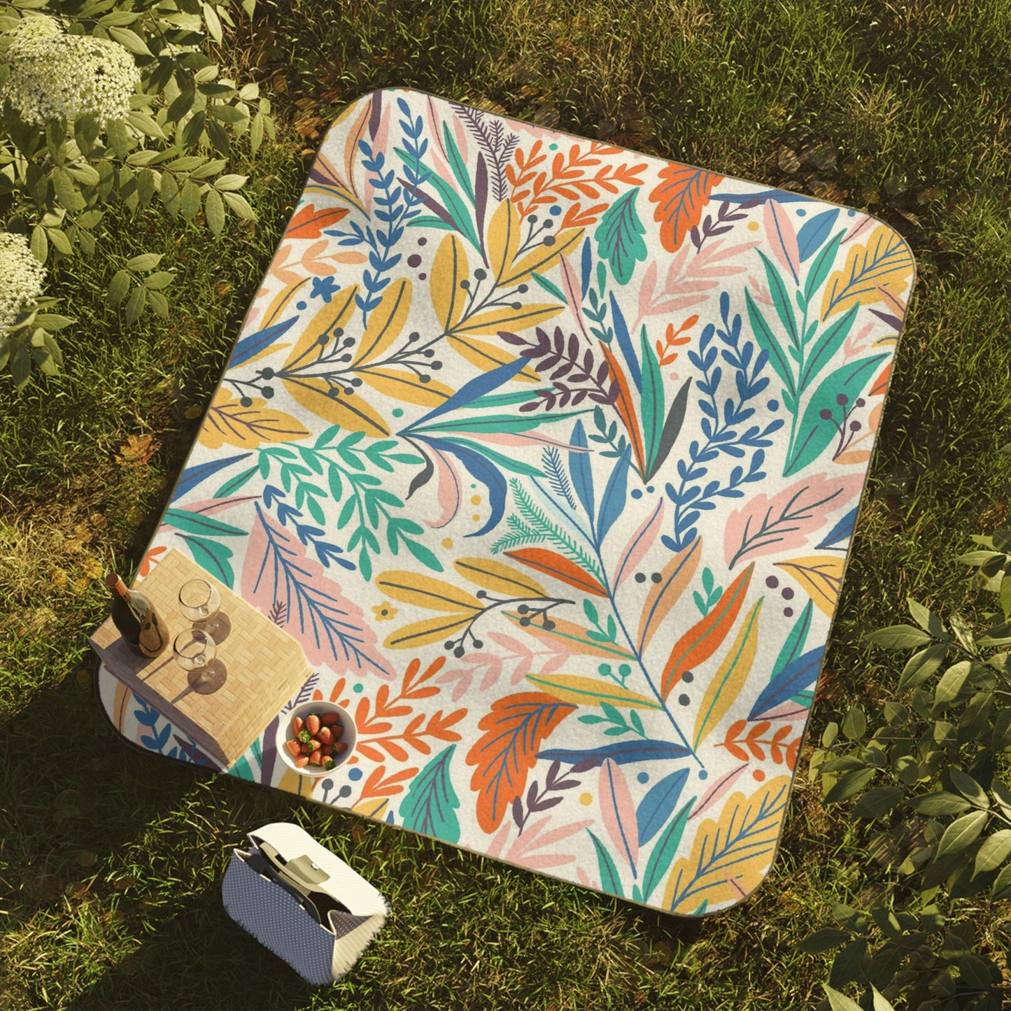 The vibrant Portable Picnic Blanket by Printify, made with water-resistant polyester and fleece, measuring 61" x 51", boasts a floral design and lies on the grass, ideal for outdoor gatherings. Nearby, a book sits next to a wooden tray holding drinks and cherries.