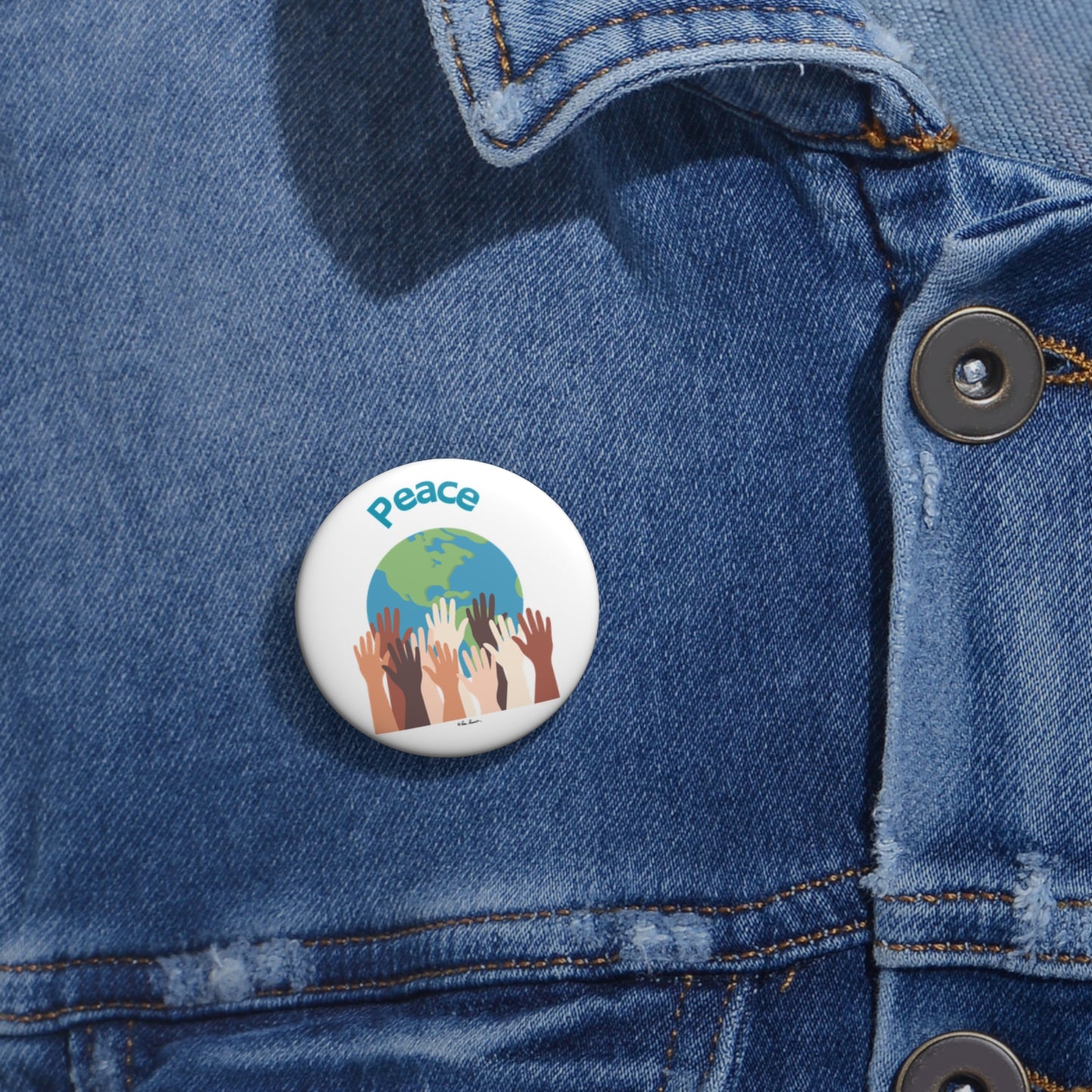 A lightweight, metal pin featuring an image of various colored hands reaching up towards a globe and the word "Peace" is attached to a denim jacket. This unisex accessory from Printify's World-Peace Buttons collection, available in three sizes, emphasizes unity and harmony.