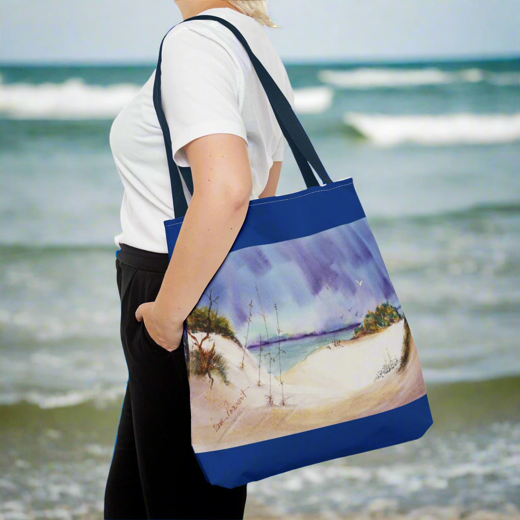 A person stands against a plain background, holding their Printify Seaside Tote Bag with a vibrant beach scene—a perfect summer adventure companion. This polyester bag is available in 3 sizes and features a 2-sided print.