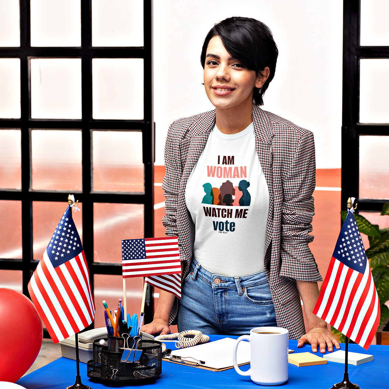 A person stands behind a desk adorned with American flags and office supplies, wearing a Printify Bella+Canvas Voting Women's T-shirt made from 100% Airlume combed and ringspun cotton. The unisex cotton jersey shirt, which reads "I AM WOMAN WATCH ME VOTE," exudes both comfort and empowerment.