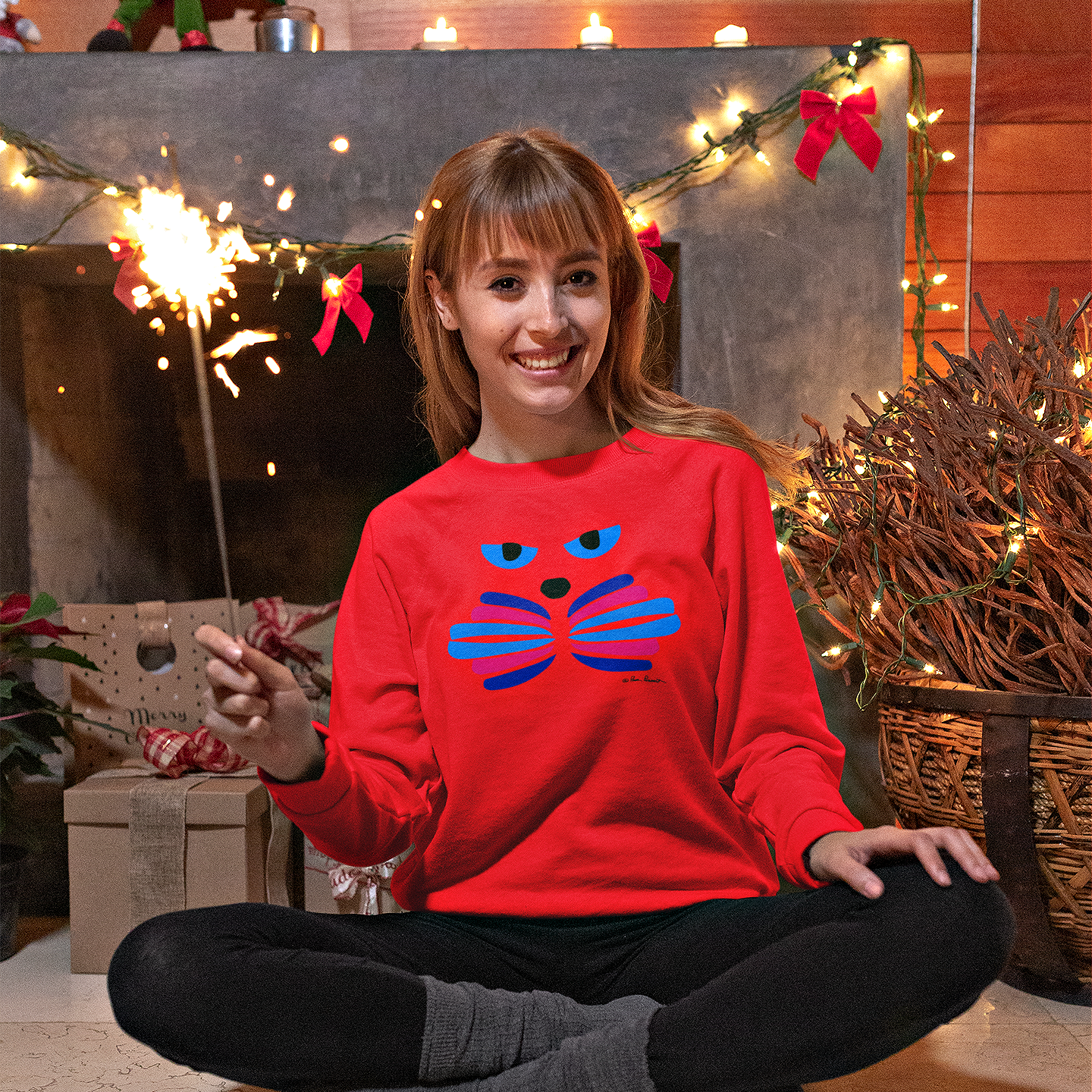 Wearing a Printify Unisex Cat-Art Sweatshirt, which is proudly made in America and comes in three color options, a woman holds a sparkler while sitting cross-legged on the floor in front of a festively decorated fireplace.