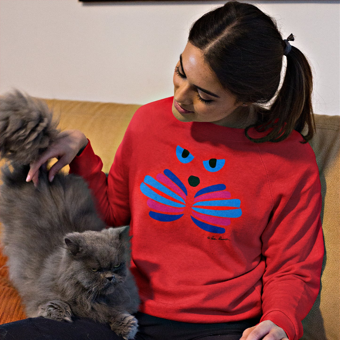 A woman wearing a blue abstract design Printify Unisex Cat-Art Sweatshirt, made from a cotton blend and available in 3 colors, sits on a couch petting a gray cat.
