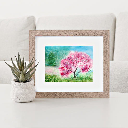 Mock up of our Pink Watercolor Print depicting a pink tree in full bloom in a meadow surrounded by a white mat and a brown wood frame (frame not included)