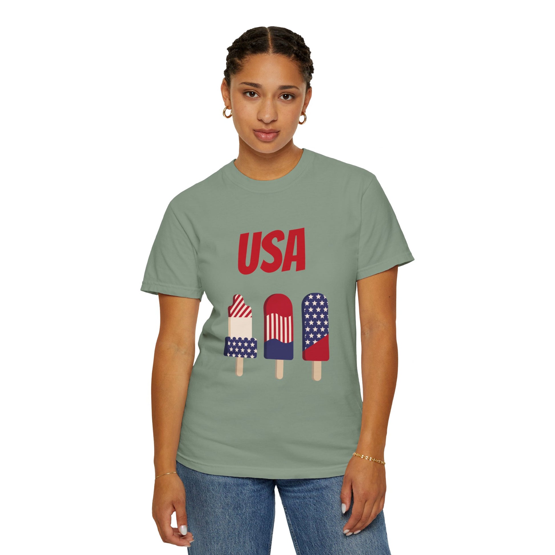 A person wearing a light green, Unisex USA T-shirt: 3 colors; Cotton; Comfort Colors 1717 by Printify made from 100% ring-spun cotton, featuring three American flag-themed popsicles printed on the front, stands against a plain backdrop.