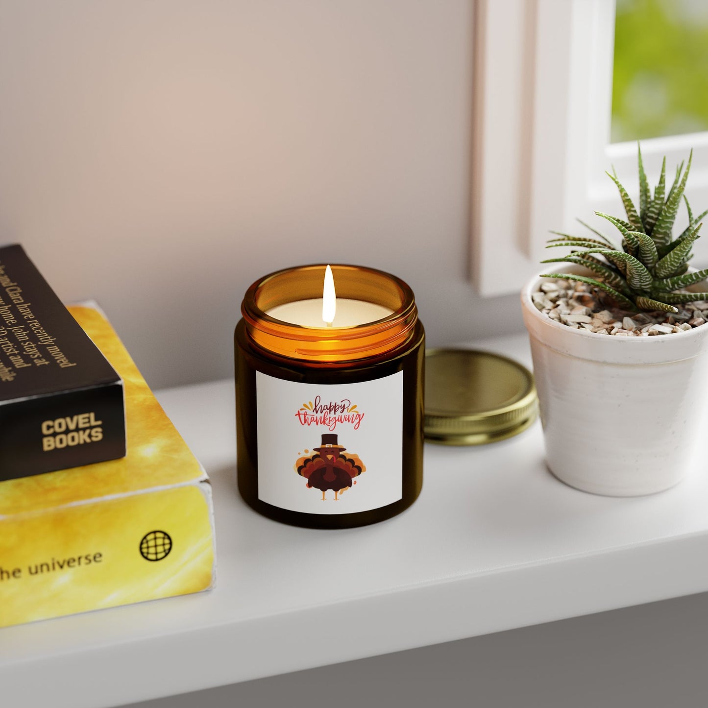 A lit Printify Thanksgiving Scented Candle (4 oz., 20 hrs, eco-friendly) in a brown glass jar sits on a white windowsill beside a potted succulent and books, filling the space with its cozy aroma.