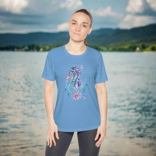 A woman in a blue Moisture-wicking Ladies T-shirt: Seashorse with a colorful design and black leggings stands by a large body of water with mountains in the background. This Printify Sport Tek 350 shirt offers both comfort and a feminine fit for active adventures.