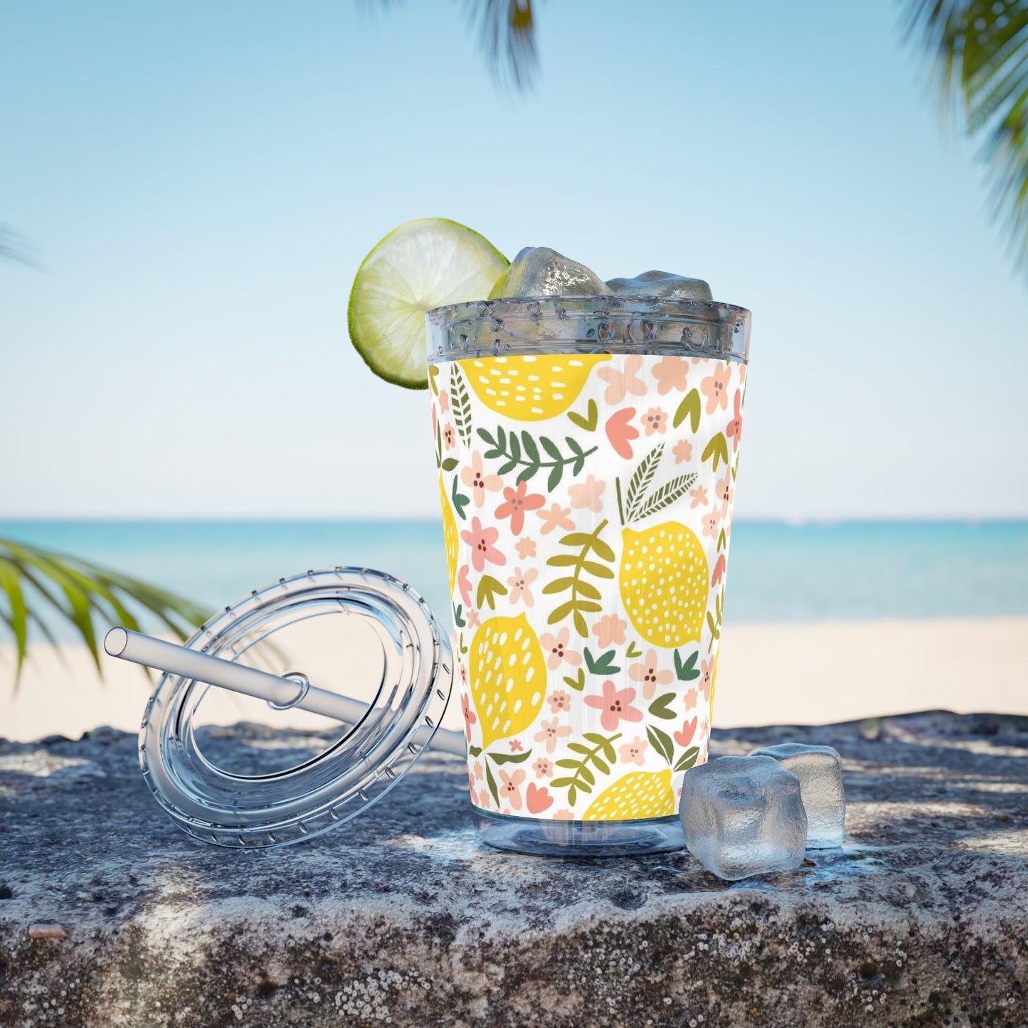 A glass of pink lemonade in a Printify Pink Lemons Tumbler on the beach with a lime wedge.
