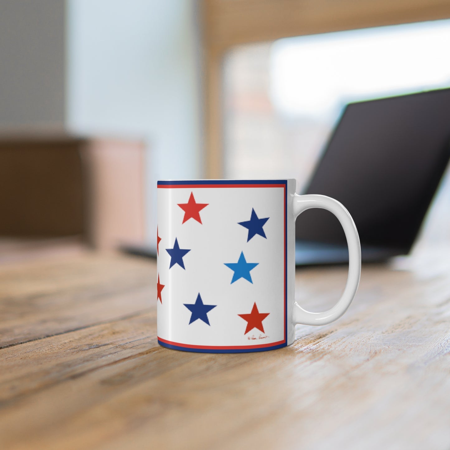 A Printify Mug with Stars: Blue and Red on White; Ceramic; 11 oz. sits on a wooden table, embodying a patriotic spirit. In the blurred background, a partially open laptop adds a modern touch to the cozy scene.