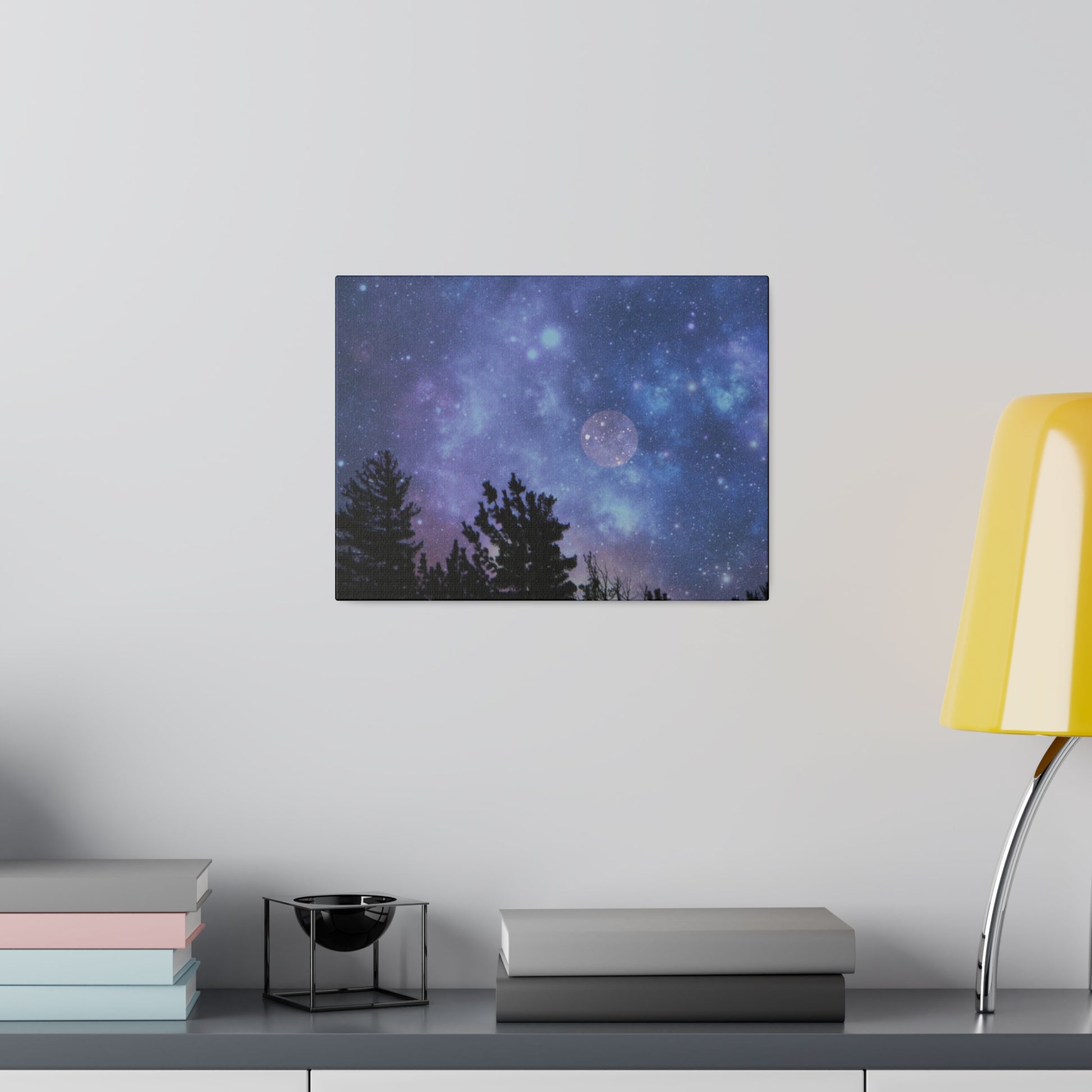 A Printify Blue-Moon Matte Canvas with a cosmic scene hangs on a wall above a stack of books next to a yellow lamp.