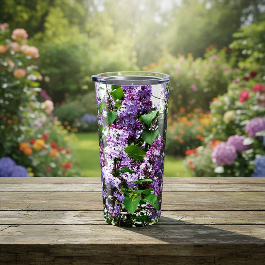 The Printify Purple Lilacs Tumbler (20 oz., stainless steel, insulated) sits on a wooden table outdoors, its clear design adorned with lilac flowers, against a vibrant garden blooming in the background.