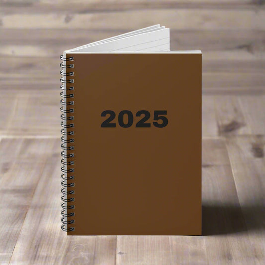 A Printify Stylish 2025 Spiral Notebook, featuring a brown cover with "2025" in bold black text, sits open on a wooden surface. This compact 6x8 notebook includes ruled lines and has 118 pages.