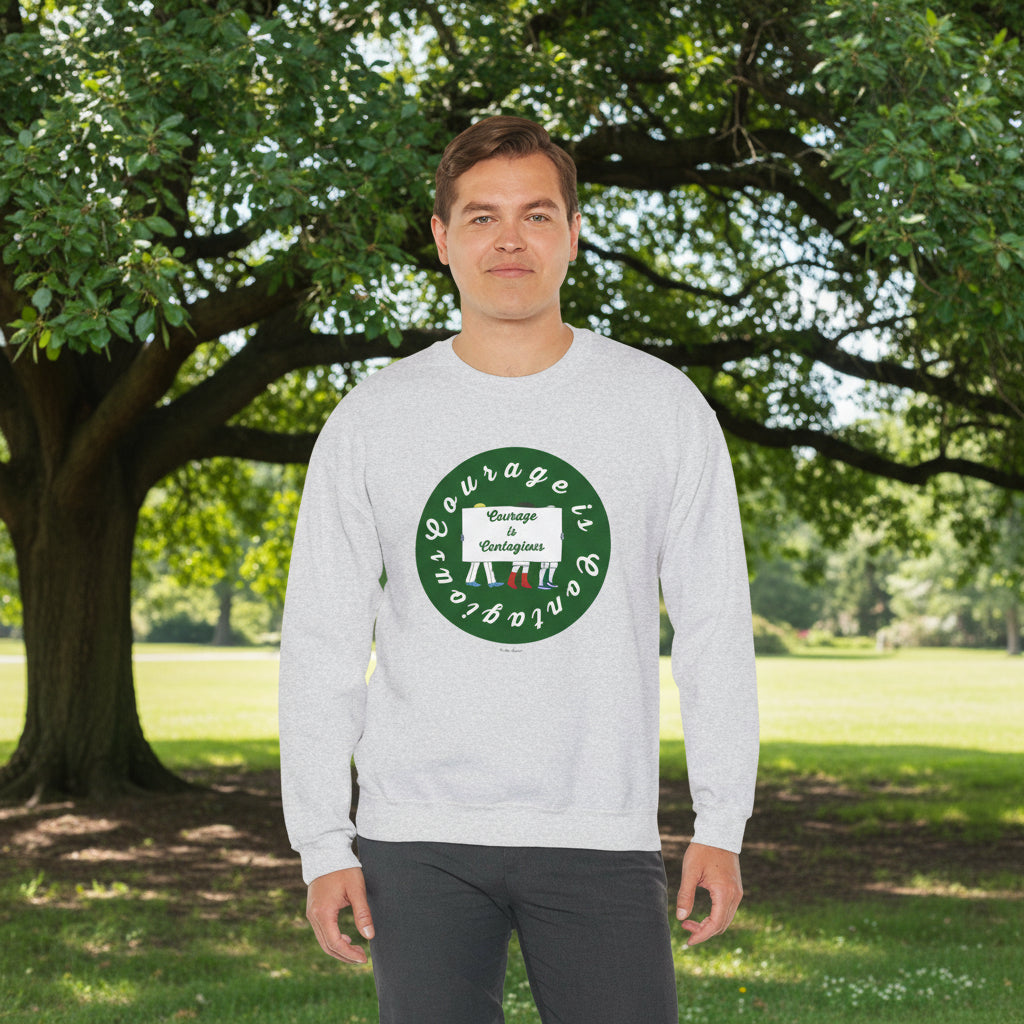 A man stands on grass before a large tree, wearing the Printify "Courage is Contagious | Protest" unisex crewneck sweatshirt, light gray with a green circular design and white text.