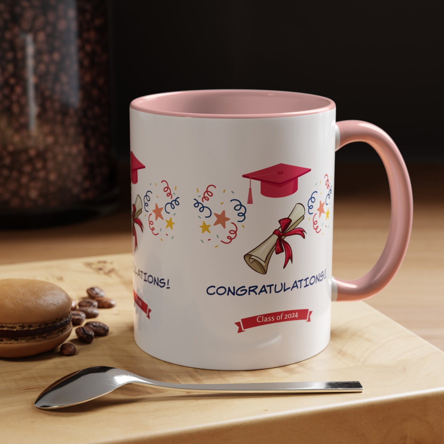A white Printify 2024 Congratulations Mug: Graduation; 11 oz.; 2 Colors with a pink handle features graduation-themed graphics, the word "Congratulations!" and "Class of 2023" alongside a spoon, coffee beans, and a macaron on a wooden surface—an ideal custom mug to celebrate milestones.