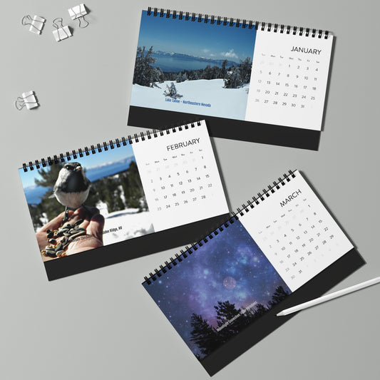 Three desk calendars from Printify's 2025 Inspirational Desktop Calendar series showcase Pam Ponsart photography with Sierra Nevada images—snowy landscape, a bird on a hand, and a starry night sky. Nearby are a pen and binder clips.