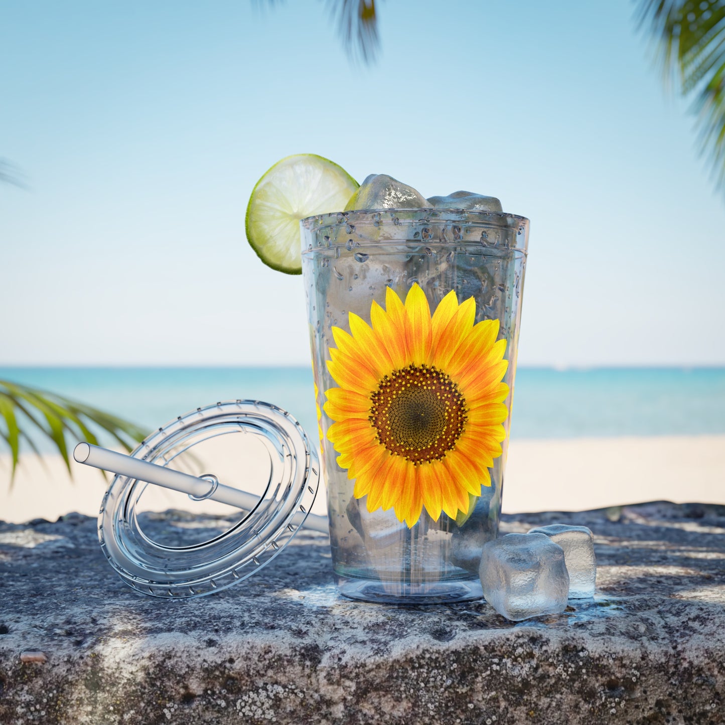 A BPA-free Acrylic Printify Golden Sunflower Tumbler with ice and a straw on a rock.