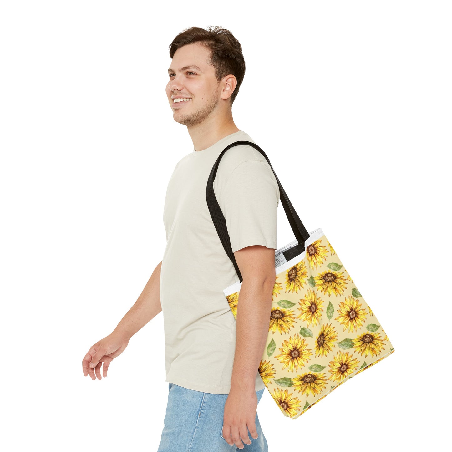 Young man smiling and walking, holding a Printify polyester tote bag with a yellow sunflower pattern, wearing a light t-shirt and jeans.