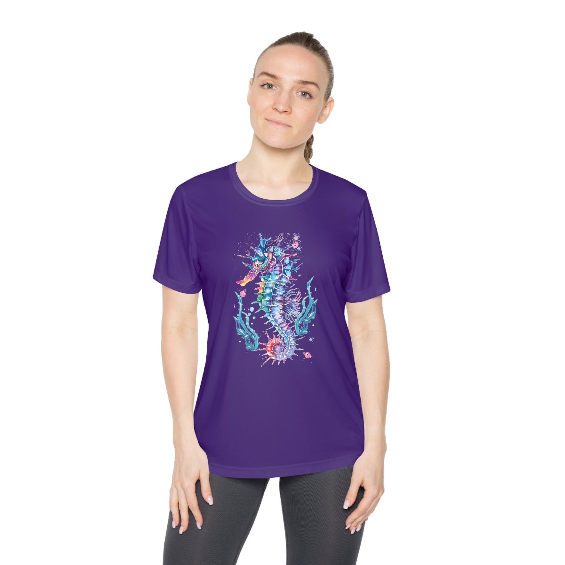 A person wearing a feminine fit, moisture-wicking Printify Moisture-wicking Ladies T-shirt: Seashorse; Sport Tek 350 in purple with a colorful seahorse design stands with a neutral expression.