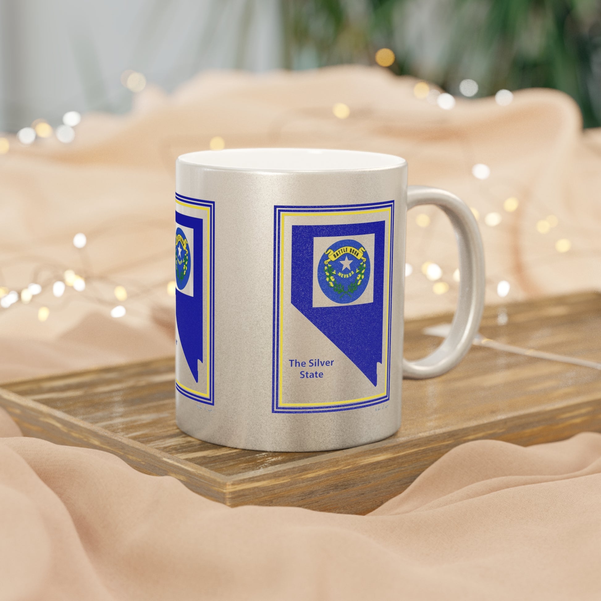 Featuring a contemporary and elegant design, the Printify Silver Metallic Mug: 11 oz., crafted from ceramic, celebrates Nevada with a blue emblem displaying "The Silver State." Ideal as a gift from Nevada, it is beautifully presented on a wooden board surrounded by soft beige fabric and illuminated string lights that create a cozy ambiance.