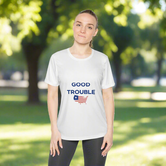 A woman stands outside wearing the Printify Ladies Good Trouble T-shirt, a short-sleeve, moisture-wicking top featuring "GOOD TROUBLE" above a U.S. flag graphic, showing her support for activism amid sunlit trees.