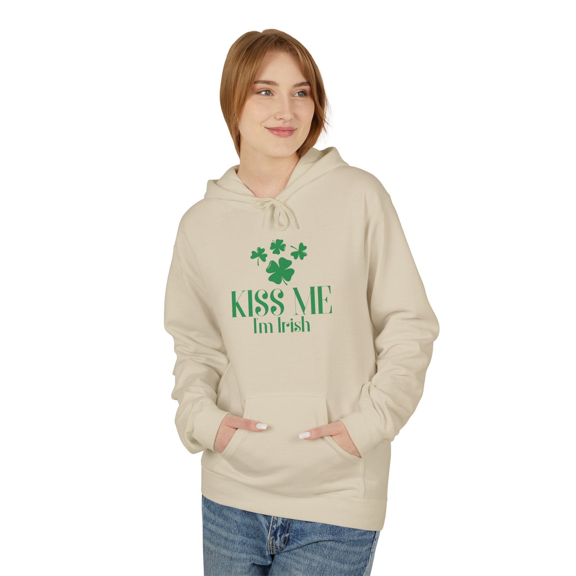 A woman wears the cream "Kiss-Me I'm Irish Hoodie: St. Patricks Day Shamrock Pullover" by Printify, featuring green text and shamrocks, with her hands in the front pocket.