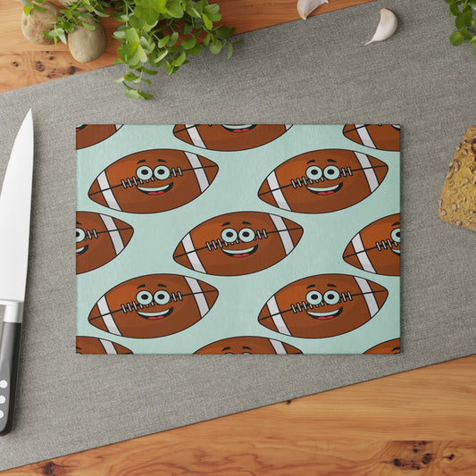 The Playful Football-Faces Cutting Board: Glass by Printify features smiling cartoon footballs on light blue, adding fun to your kitchen decor; available in 2 sizes and shown with a knife, bread, and greens on the counter.