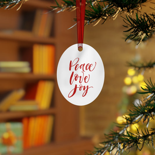 A Holiday-Text Metal Ornament from Printify hangs on the Christmas tree branch, featuring an oval shape and a red hanging ribbon, bringing festive charm to your holiday decor. The ornament is adorned with "Peace Love Joy" in red script. A blurred bookshelf in the background enhances the elegance of this Christmas tree decoration.