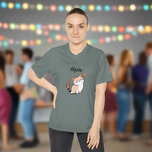 A person wears a Printify Women's Tabby-Cat T-shirt: V-neck; UV 40+; Team 365 featuring a cartoon cat and the word "Meow" printed on it, made of moisture-wicking polyester, standing in front of a blurred background with people and colorful string lights.