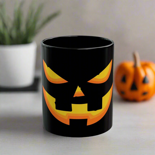 The Printify Halloween Pumpkin Face Mug, a black ceramic mug with a spooky yellow and orange jack-o'-lantern design, sits on a table with a blurred pumpkin decoration and plant in the background.