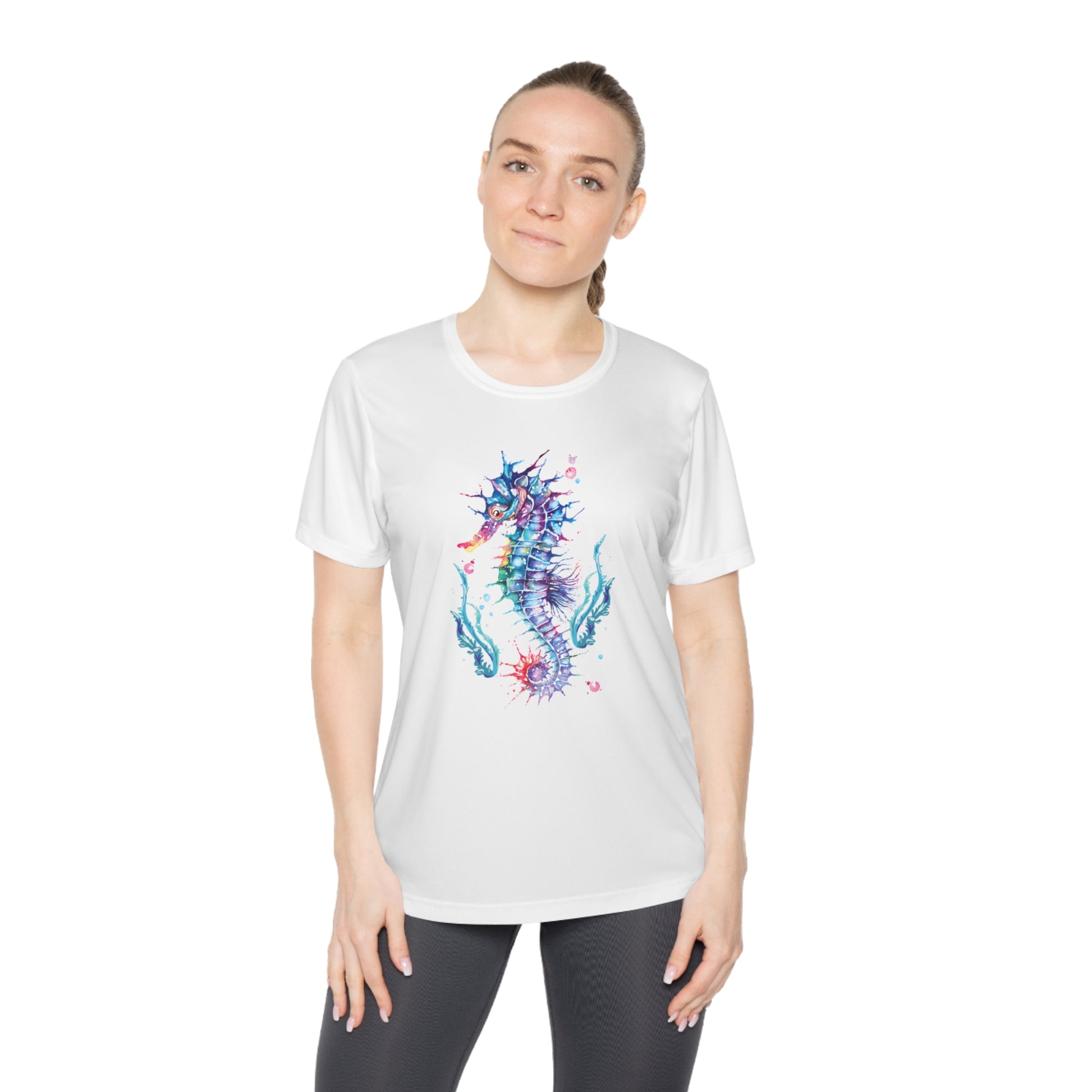 A person standing and wearing a Printify Sport Tek 350 Moisture-wicking Ladies T-shirt: Seashorse featuring a colorful seahorse design. They have long hair tied back and are dressed in black workout leggings.