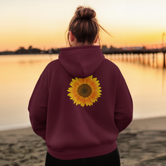 A person with a bun stands on a beach at sunset wearing the Printify Unisex Sunflower Zip Hoodie, a full-zip sweatshirt for women and men, featuring a large sunflower graphic on the back.