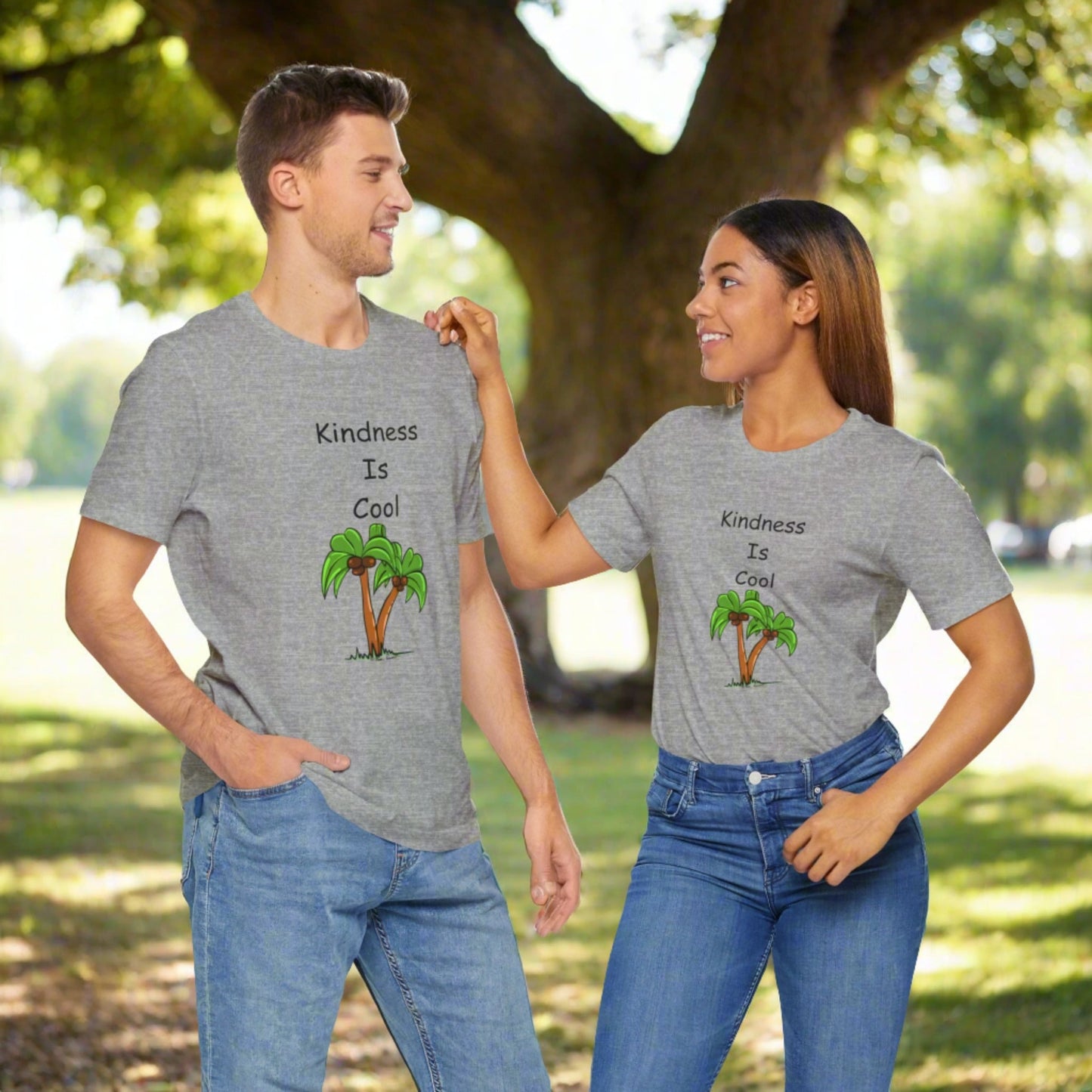 Two people in a park, wearing matching Printify Unisex Kindness-Is-Cool T-shirts made from soft cotton jersey by Bella+Canvas, stand facing each other while smiling and interacting. The grey T-shirts feature palm tree graphics and the text "Kindness-Is-Cool," enhancing their comfort as they share a moment of kindness.