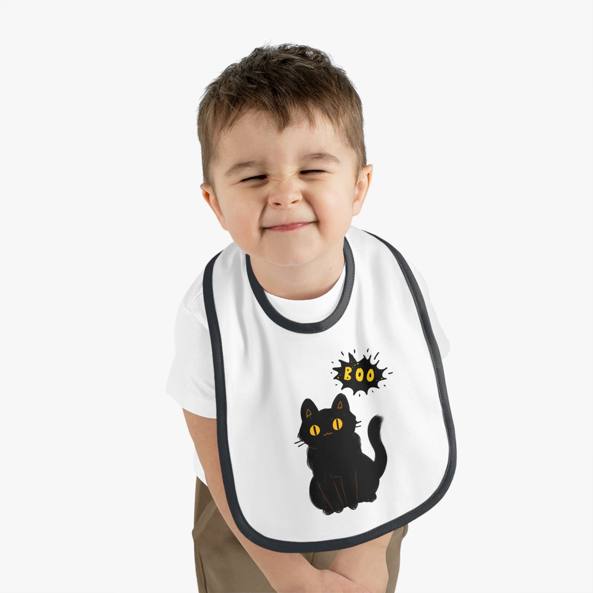 A young child smiles with eyes closed, wearing the Printify Halloween Cat Baby Bib: Cotton | Spooky Cute | Babyware—featuring a black cat and “Boo”—the perfect Halloween gift for babies.