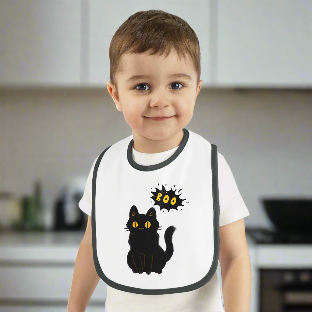 A young child stands in a kitchen wearing the Printify Halloween Cat Baby Bib: Cotton | Spooky Cute | Babyware—a white bib with a black cat and "BOO"—making it an ideal Halloween baby gift.