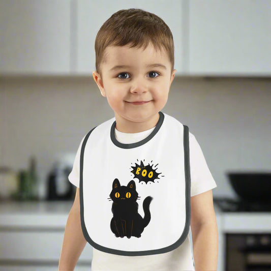 A young child stands in a kitchen wearing the Printify Halloween Cat Baby Bib: Cotton | Spooky Cute | Babyware—a white bib with a black cat and "BOO"—making it an ideal Halloween baby gift.